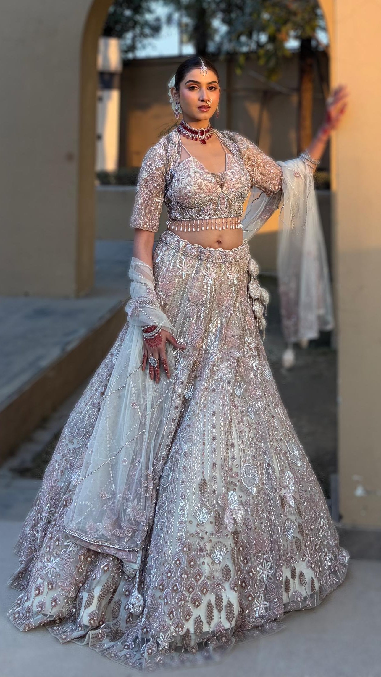 Woman in a traditional outfit standing in front of an archway with a beige wall and tree shadows.